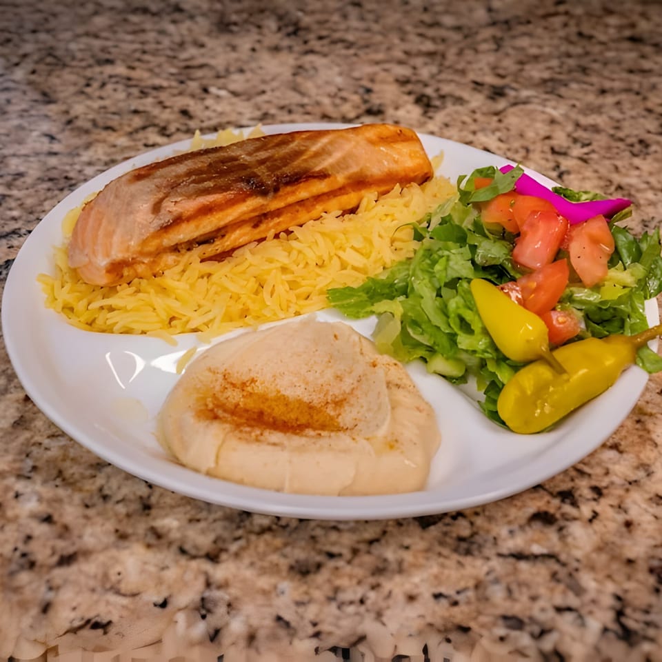 Grilled salmon with yellow rice, hummus, a green salad with tomatoes, and pickled peppers.