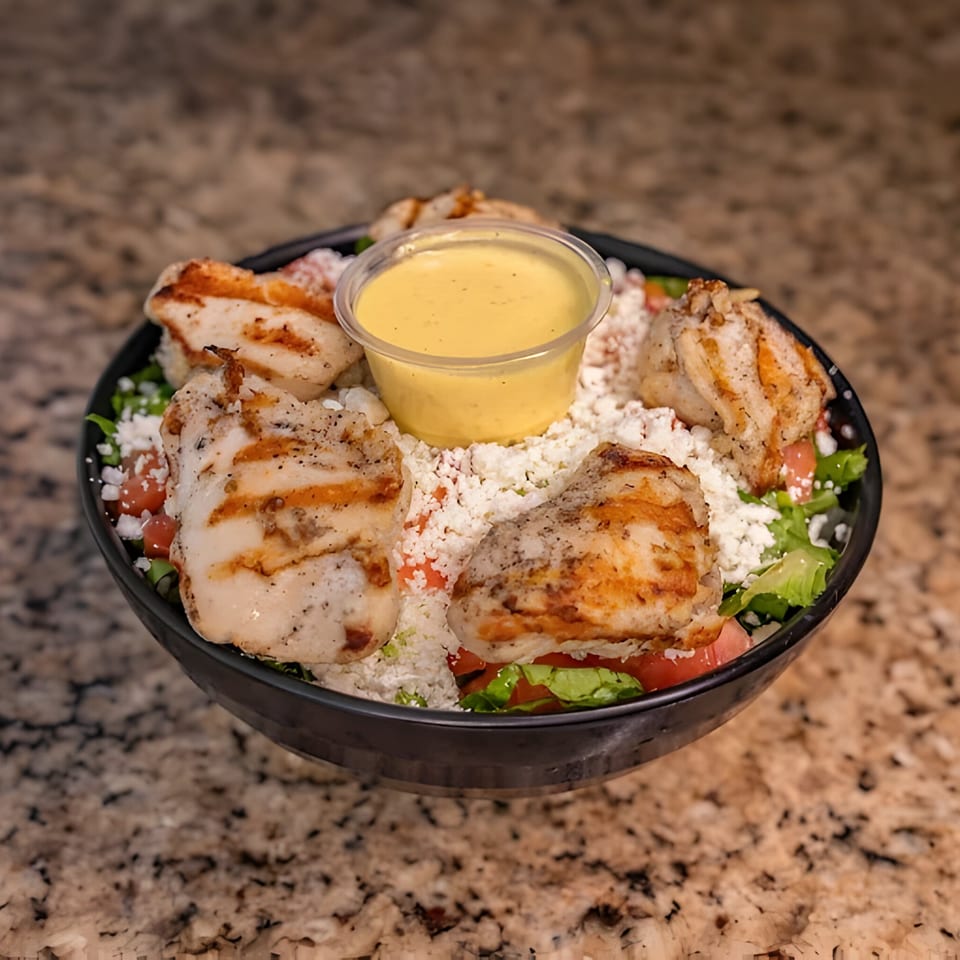 Grilled chicken pieces, crumbled feta, lettuce, and tomatoes in a black bowl with a side of dressing.